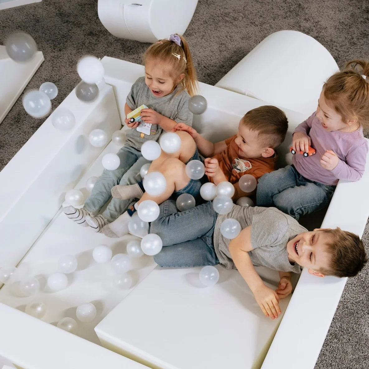Four young children playing together in white ball pit filled with soft play balls
