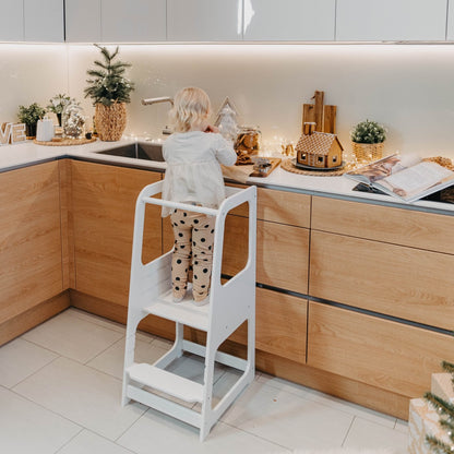 Young child standing on white Montessori helper tower step stool at kitchen counter