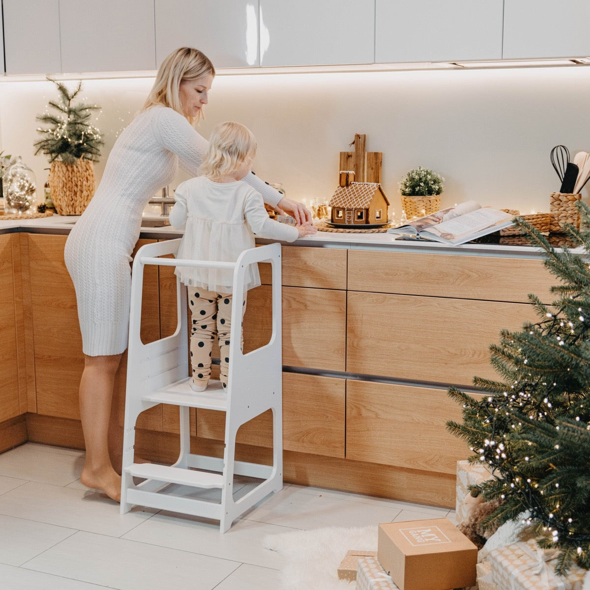 Mother assisting toddler on white helper tower stool while cooking in modern kitchen