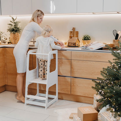 Mother assisting toddler on white helper tower stool while cooking in modern kitchen