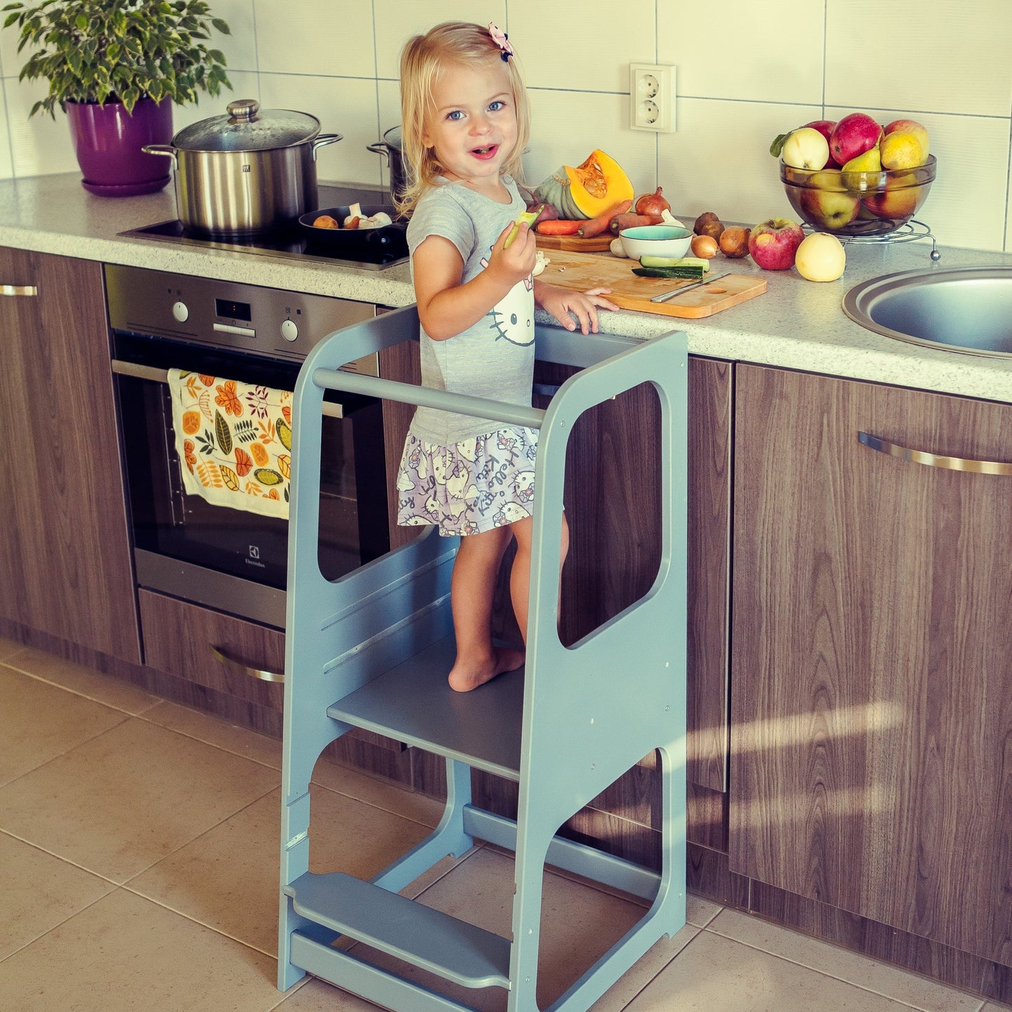 Child standing on light blue helper tower stool reaching stovetop to cook with adult supervision