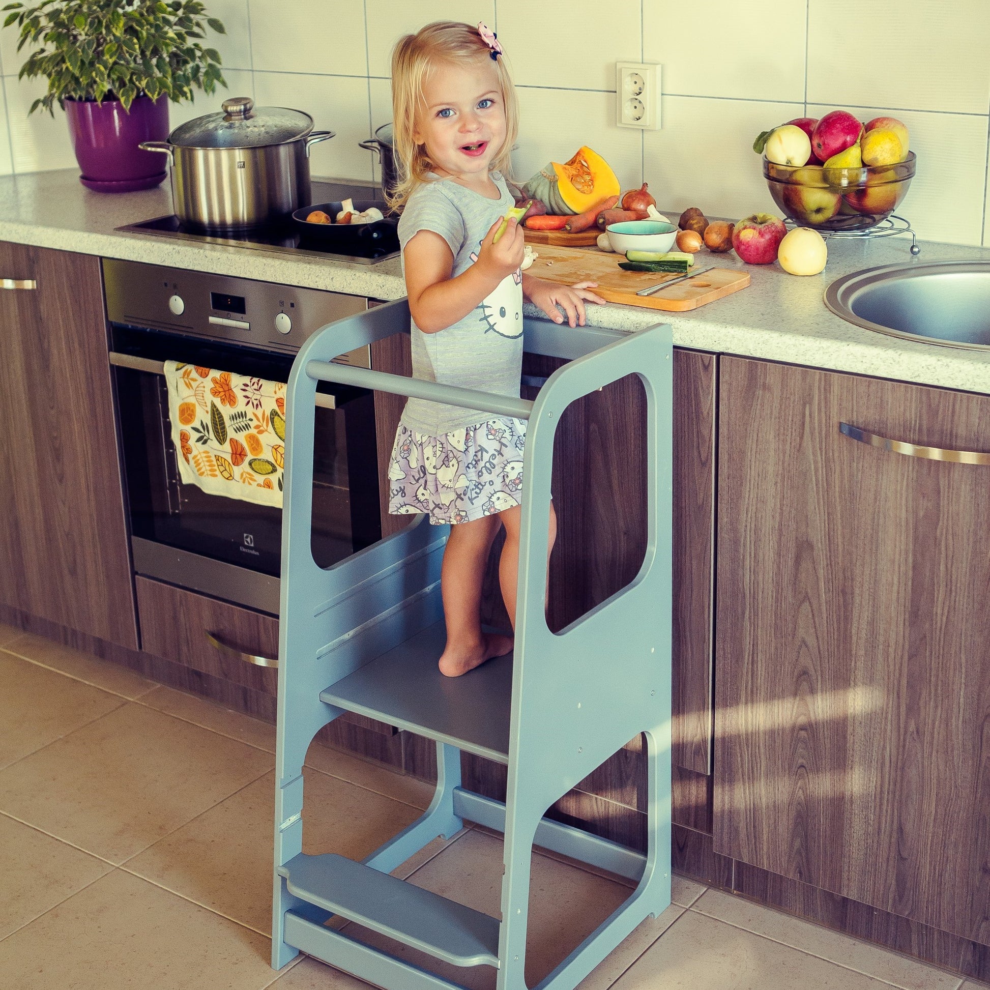 Child standing on light blue helper tower stool reaching stovetop to cook with adult supervision