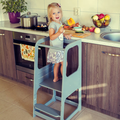 Child standing on light blue helper tower stool reaching stovetop to cook with adult supervision