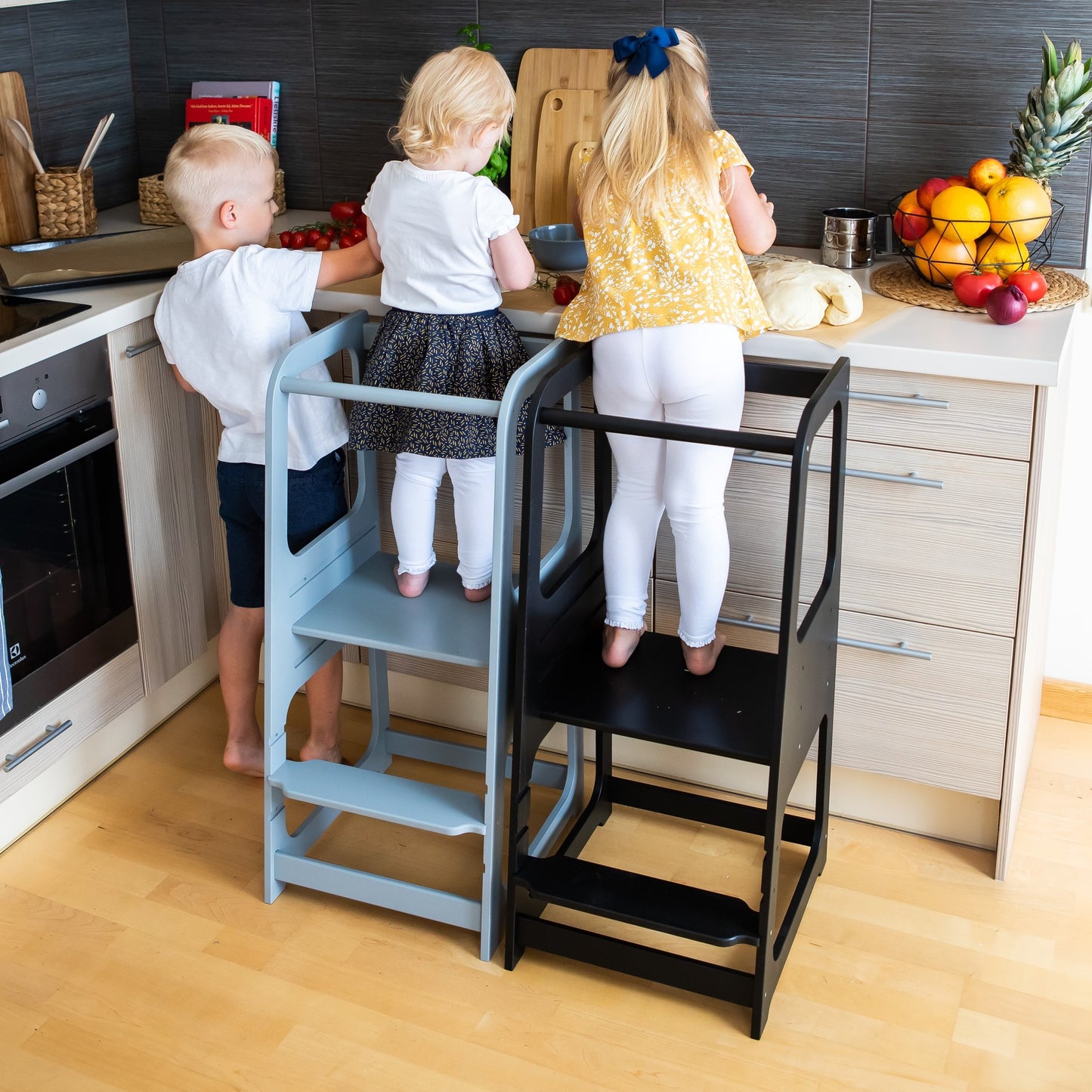 Two children cooking together using separate helper tower stools in kitchen