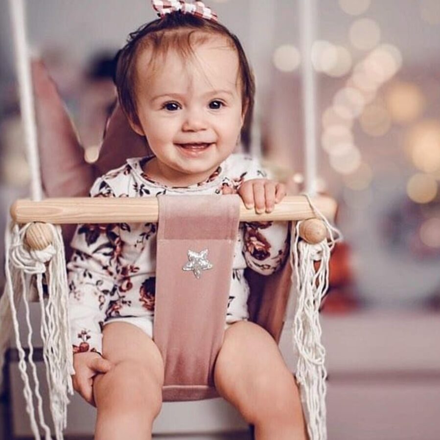 Smiling child in crown-shaped dusty pink velvet swing seat with wooden bar and white rope
