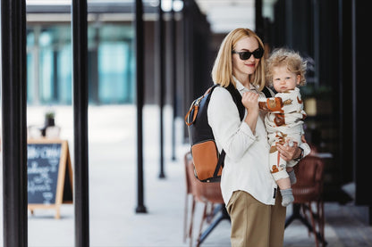 Woman wearing large black coffee diaper backpack while holding toddler outdoors