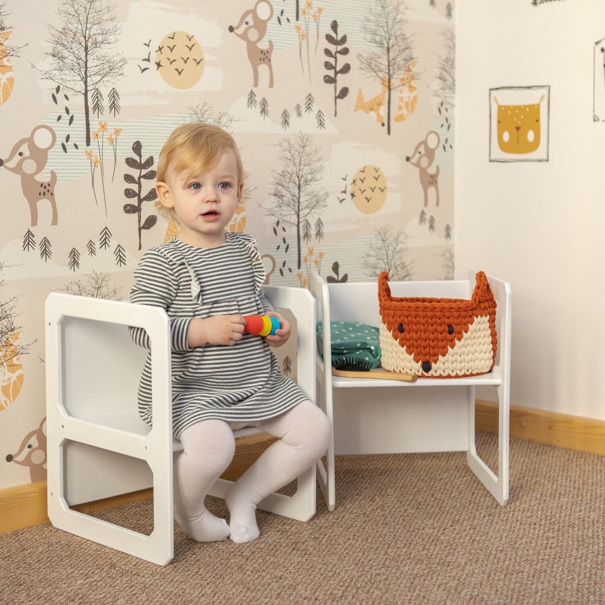 Toddler in striped shirt sitting on white Montessori chair holding toy blocks with storage table