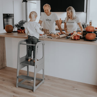Toddler standing on white learning tower at kitchen counter cooking with family