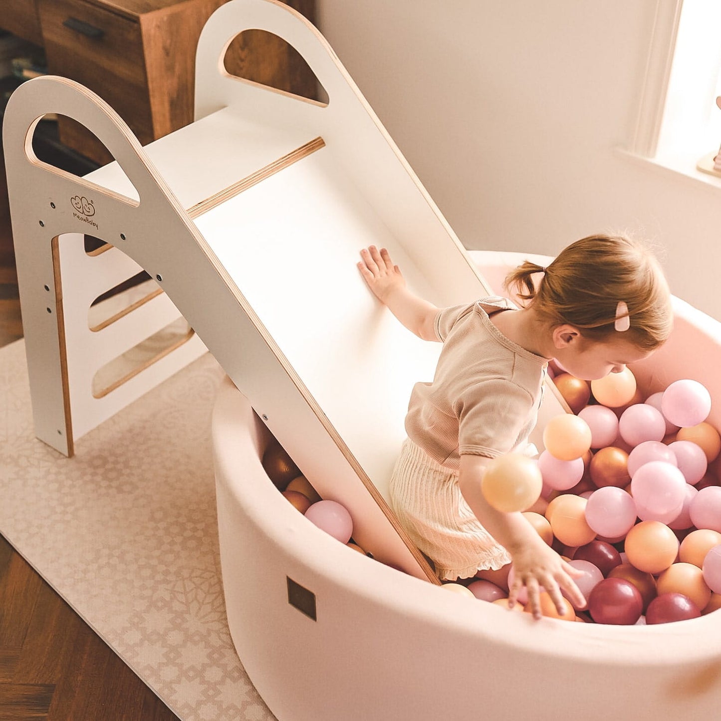 Baby sliding down white wooden play slide above pink ball pit basin