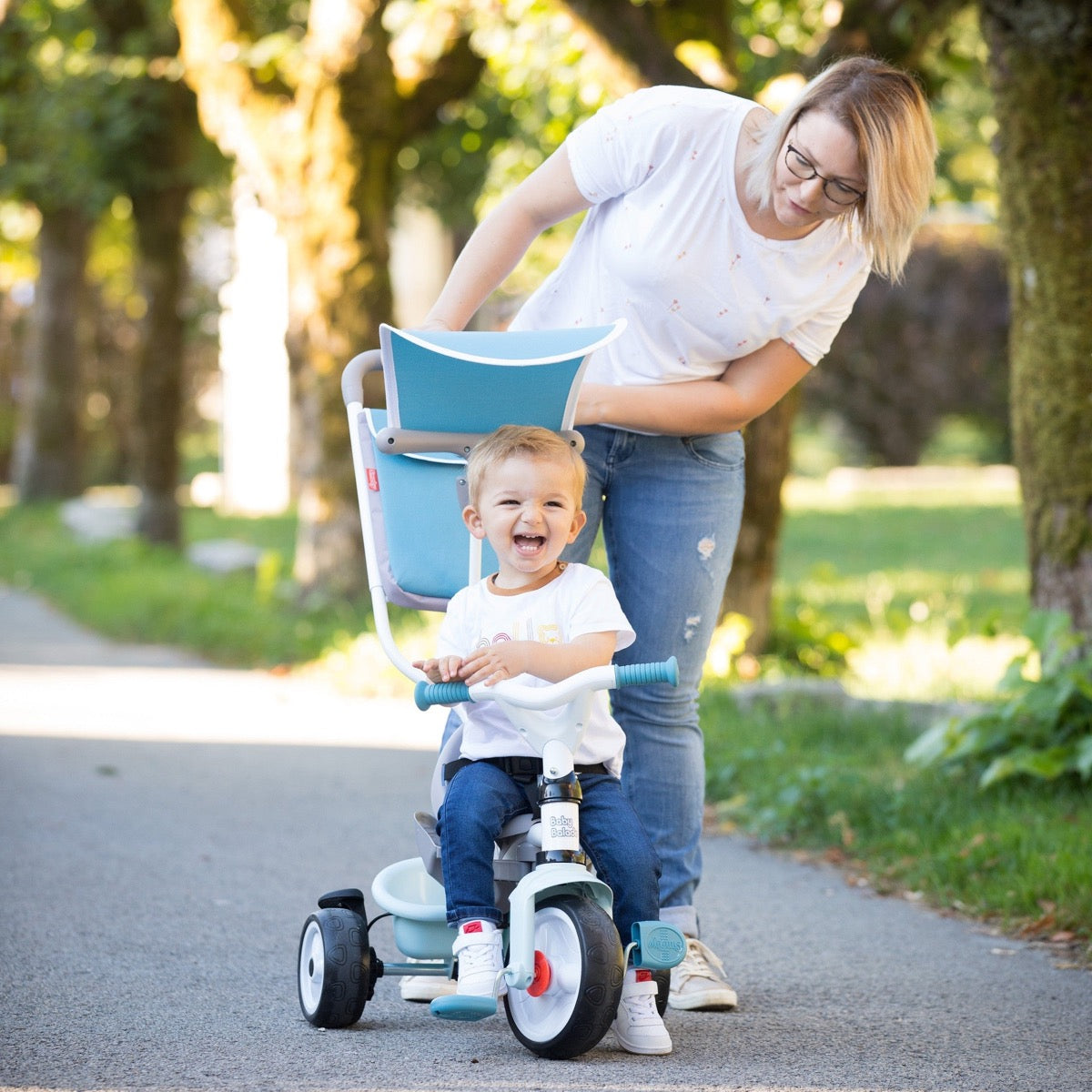 Parent pushing young child on blue Baby Balade tricycle with adjustable handle during outdoor play