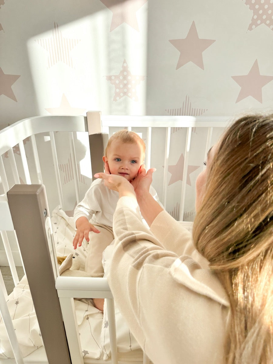 Mother interacting with baby inside white ELIN crib with star-patterned nursery wall