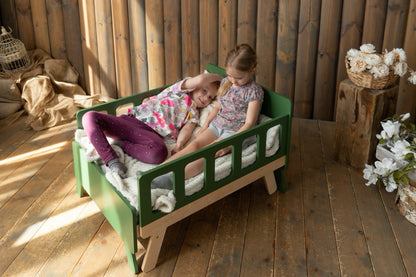 Two sisters cuddling on green Family-scl growing bed in sofa mode – compact kids bed for small rooms
