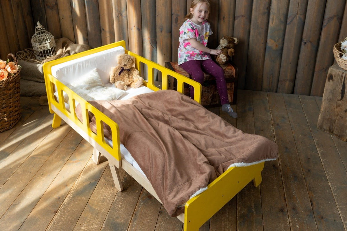 Girl sitting behind yellow Family-scl kids growing bed with teddy bear – wooden children's bed in rustic room