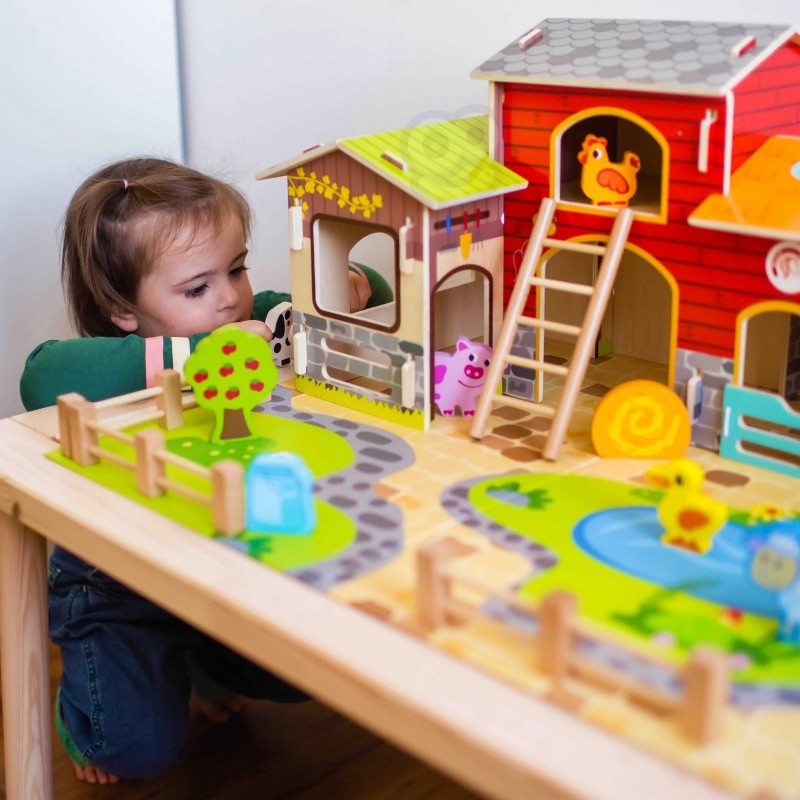 Toddler examining wooden farm buildings and animal pieces on natural wood table during creative play