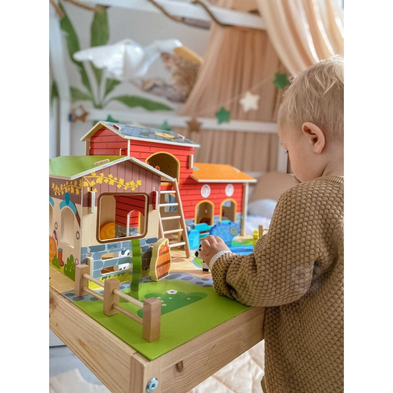 Toddler engaging with wooden farm set on table showing stable, barn, and blue water feature details