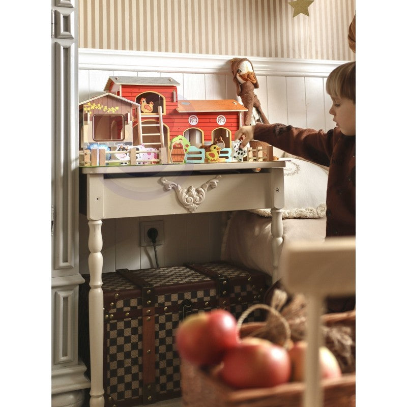 Child's hand reaching toward wooden farm set on white table with barn structures and animal toys