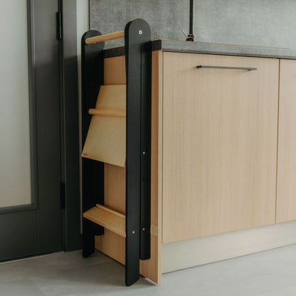 Folded kitchen tower with black frame and natural wood steps stored compactly next to kitchen cabinet