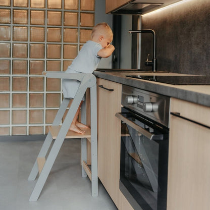 Toddler in blue outfit standing on kitchen tower at counter height reaching toward kitchen sink and faucet