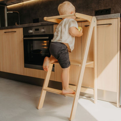 Young child climbing onto natural wood kitchen tower with white frame and safety rail in modern kitchen