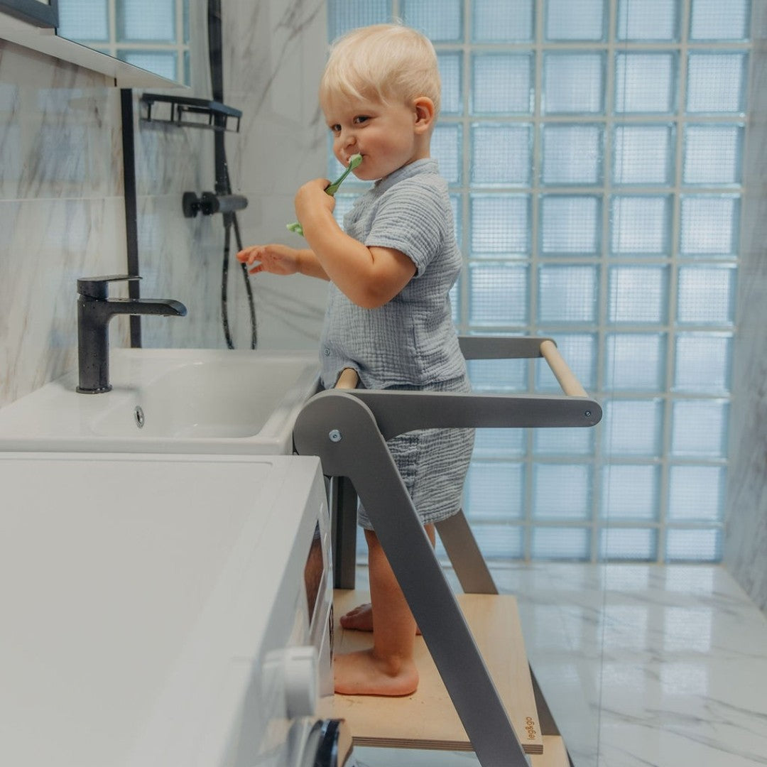 Toddler in grey shirt reaching countertop while standing on natural wood kitchen tower with safety features