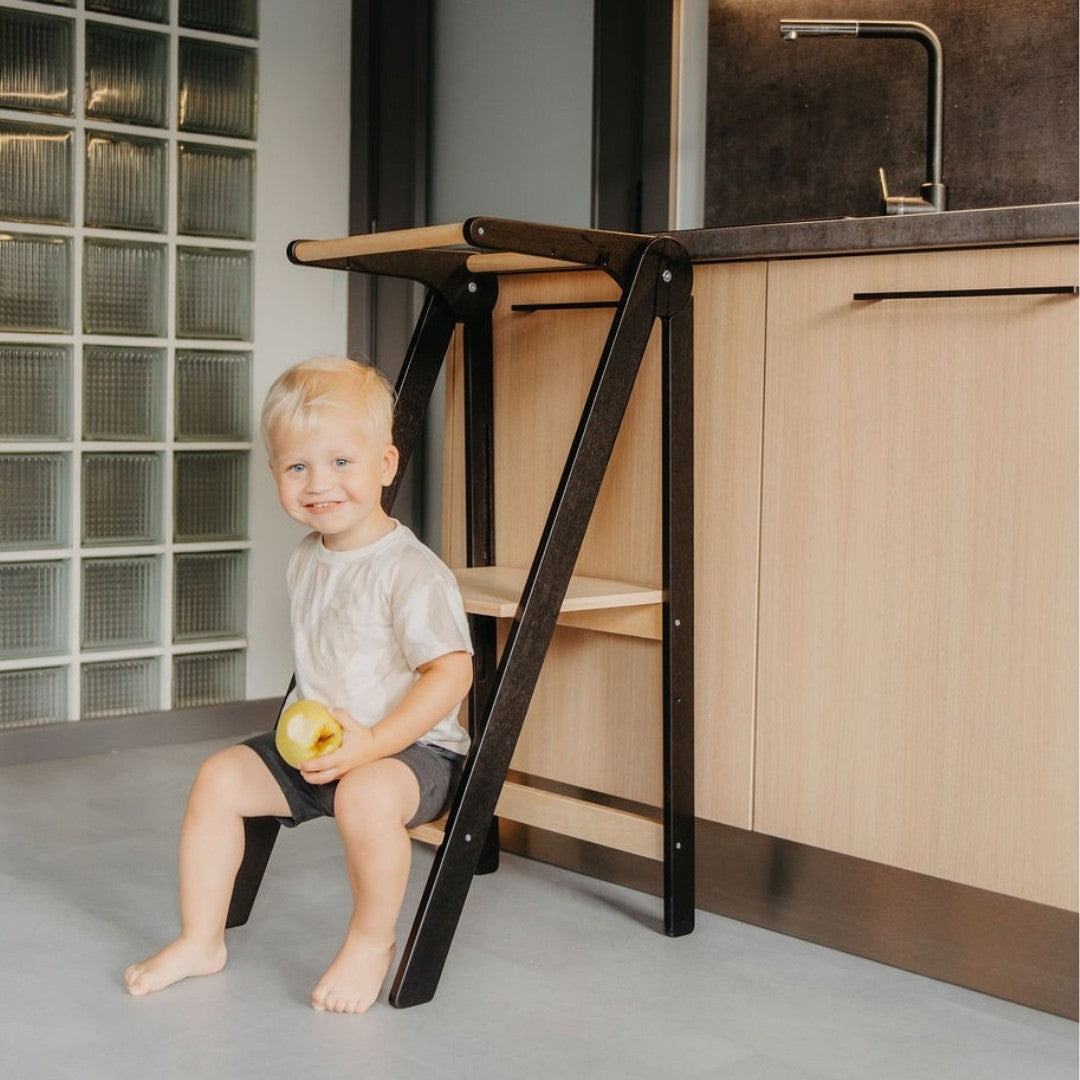 Toddler with apple sitting on black kitchen tower platform at dining table showing countertop accessibility