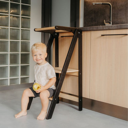 Toddler with apple sitting on black kitchen tower platform at dining table showing countertop accessibility