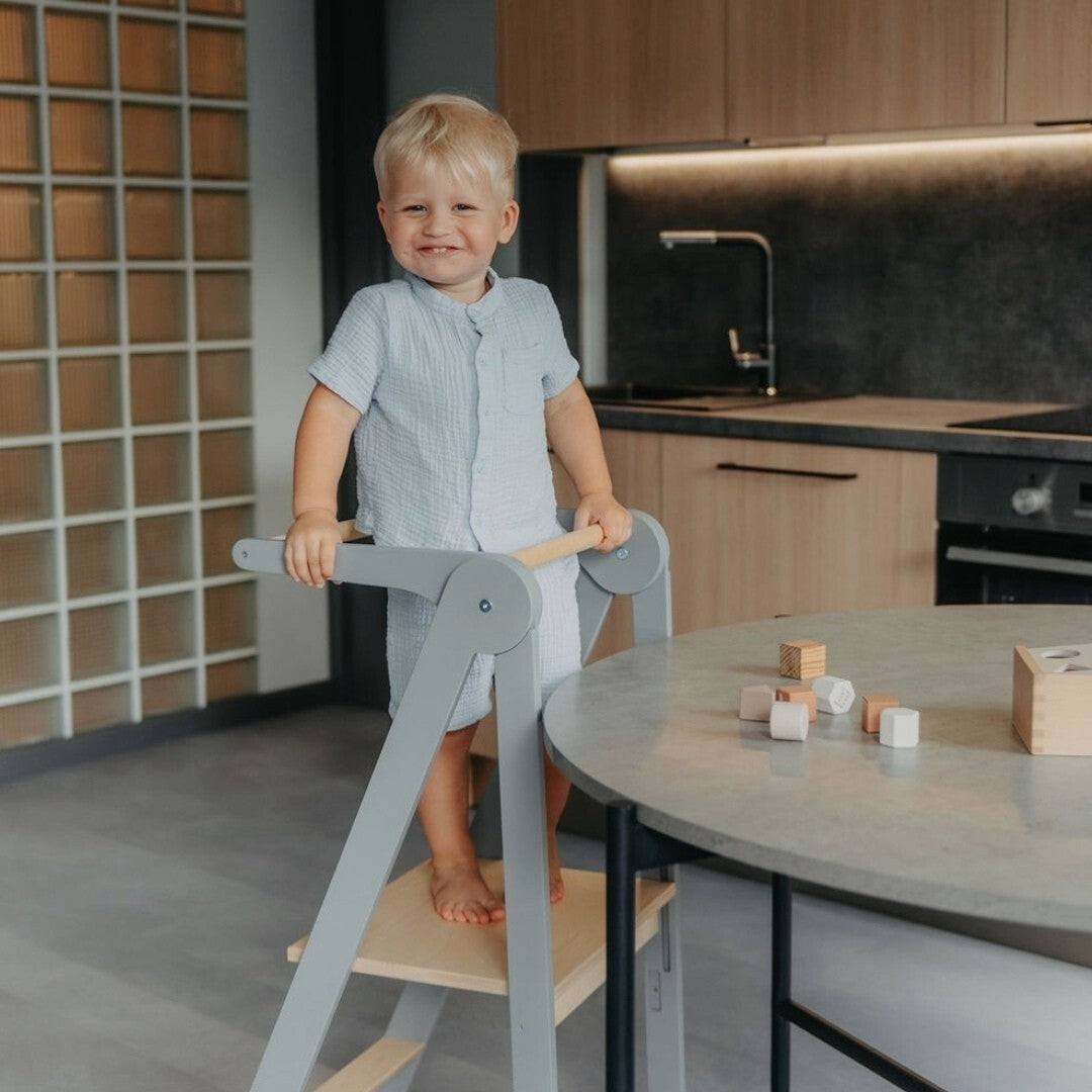 Small child in light blue shirt using removable rail on grey kitchen tower at tall table in kitchen