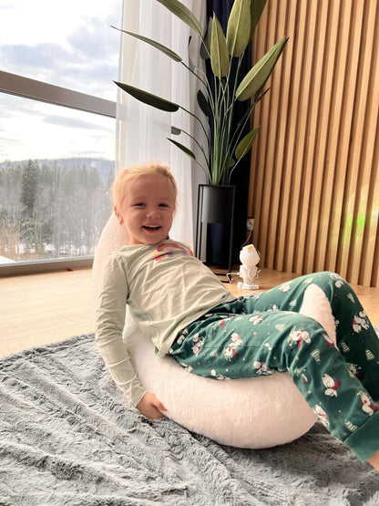 Young child sitting on white half-moon pillow by window, enjoying cozy playtime