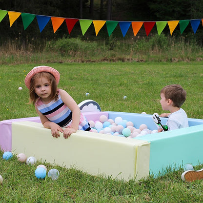 Two children playing in light pastel foam ball pit outdoors on grass with colorful bunting decoration
