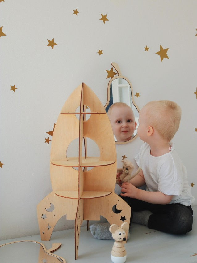 Toddler looking at pear mirror alongside wooden rocket shelf in playroom with star wall decals