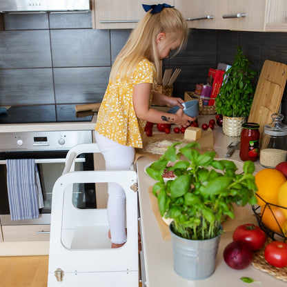 Young child standing on white kitchen tower reaching counter while cooking with adult