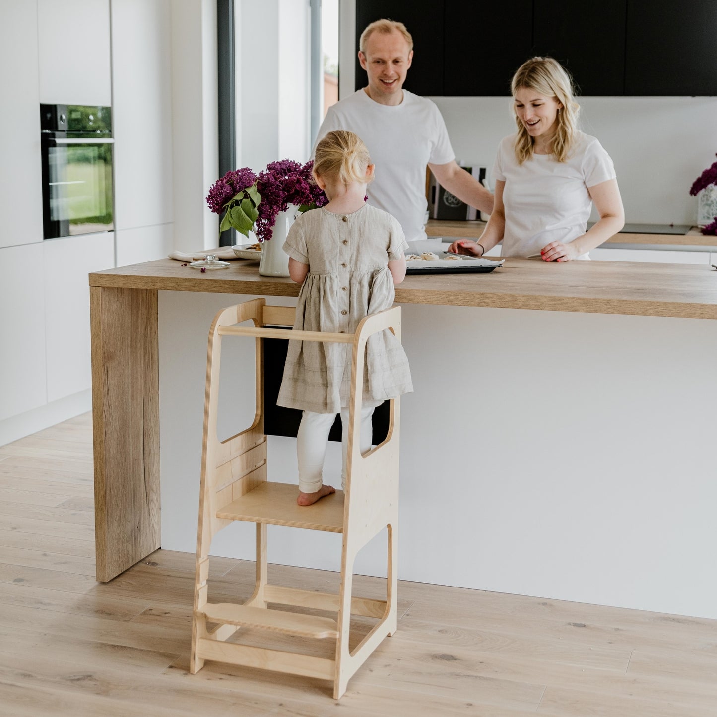 Child standing on natural wood Montessori kitchen tower at counter height with parents