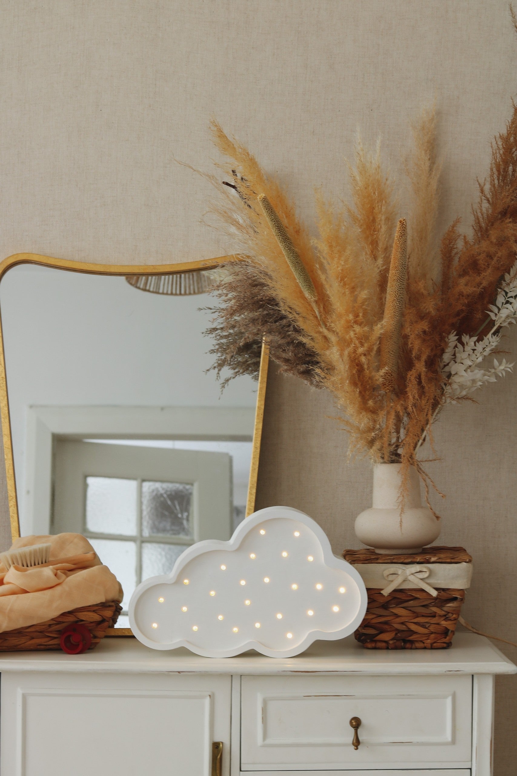 White cloud lamp glowing on dresser beside gold mirror and dried pampas grass decor