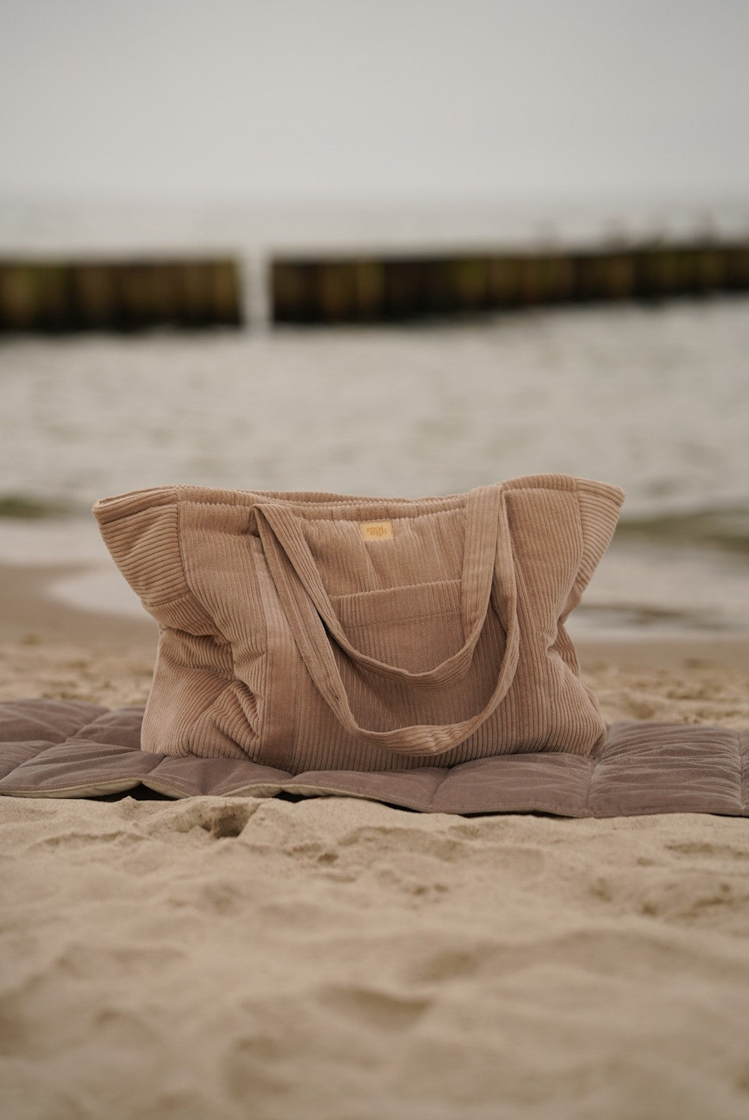 Latte quilted weekender bag sitting on sand with ocean backdrop