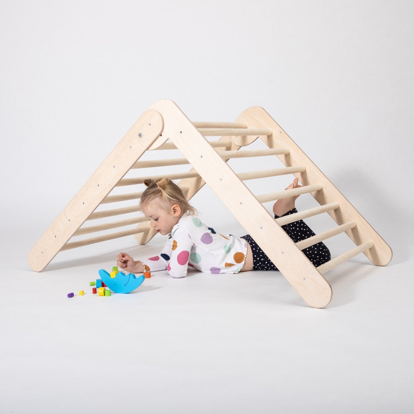 Child climbing wooden triangle with toy basket and colorful toys on floor