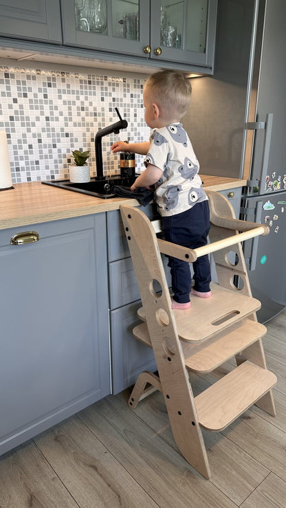 Child standing on wooden learning tower helping at kitchen counter with an adult nearby