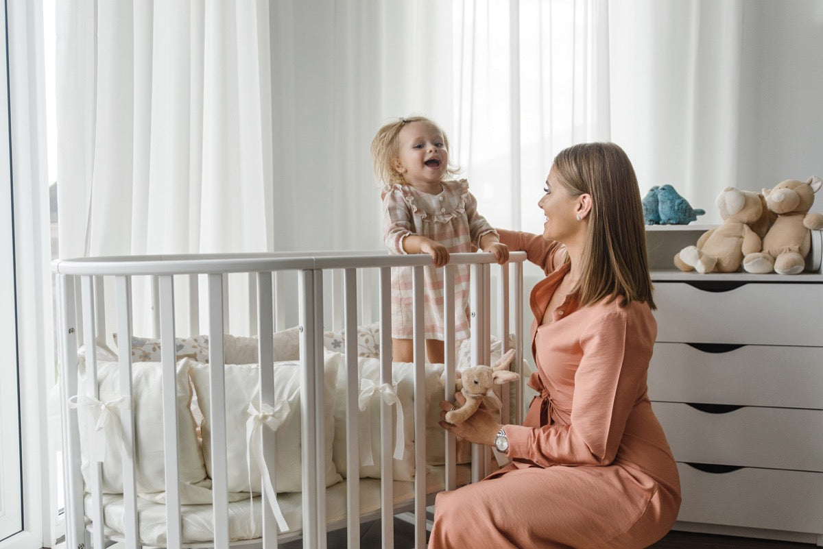 Mother lifting toddler from white wooden round crib in bright nursery