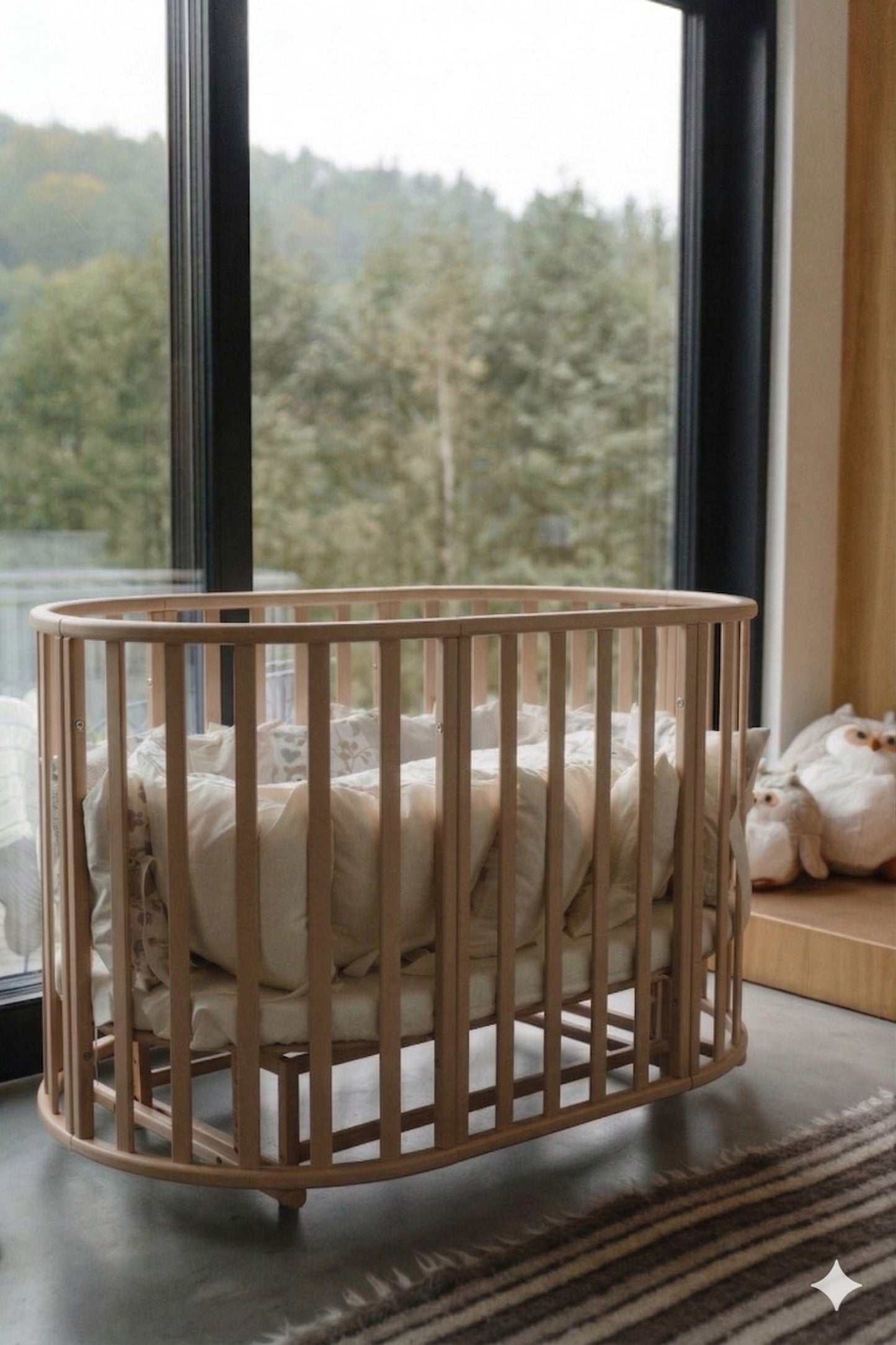Round cappuccino beechwood baby crib with mattress displayed by window with forest view