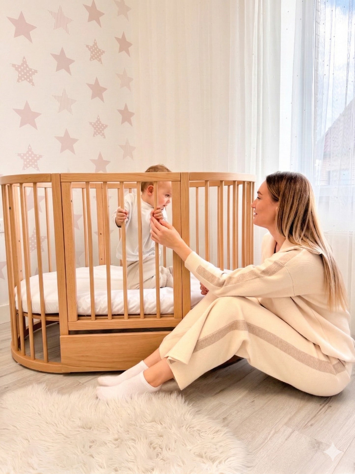 Mother sitting beside round crib with toddler inside in bright nursery