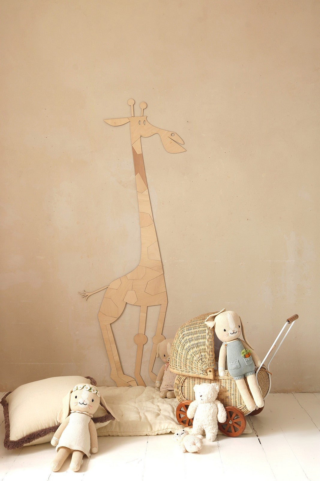 Wooden giraffe wall art standing next to wicker chair with stuffed animals and toys on white floor