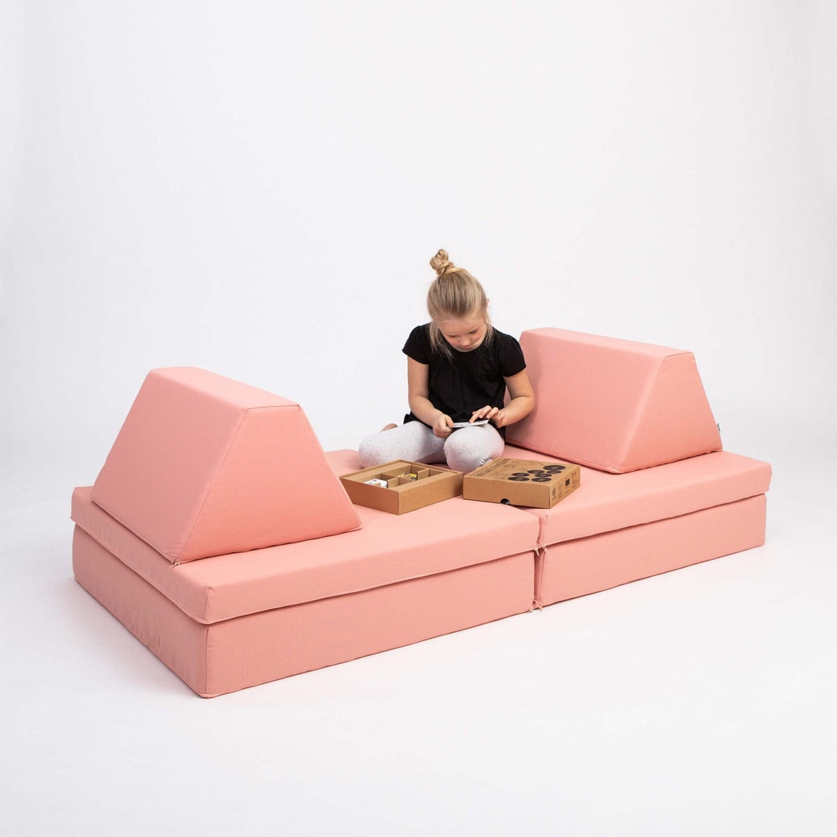 Child sitting on salmon pink foam play sofa with angled back cushions and storage baskets