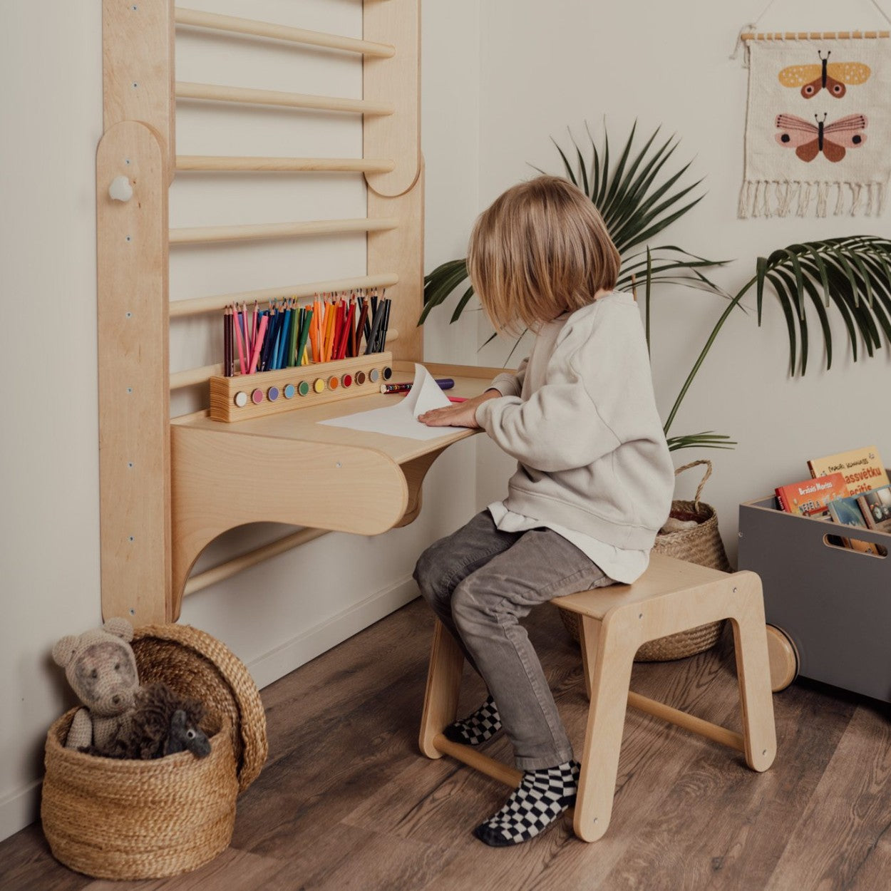 Child sitting at wooden desk attached to Swedish wall, using colored pencils and stool