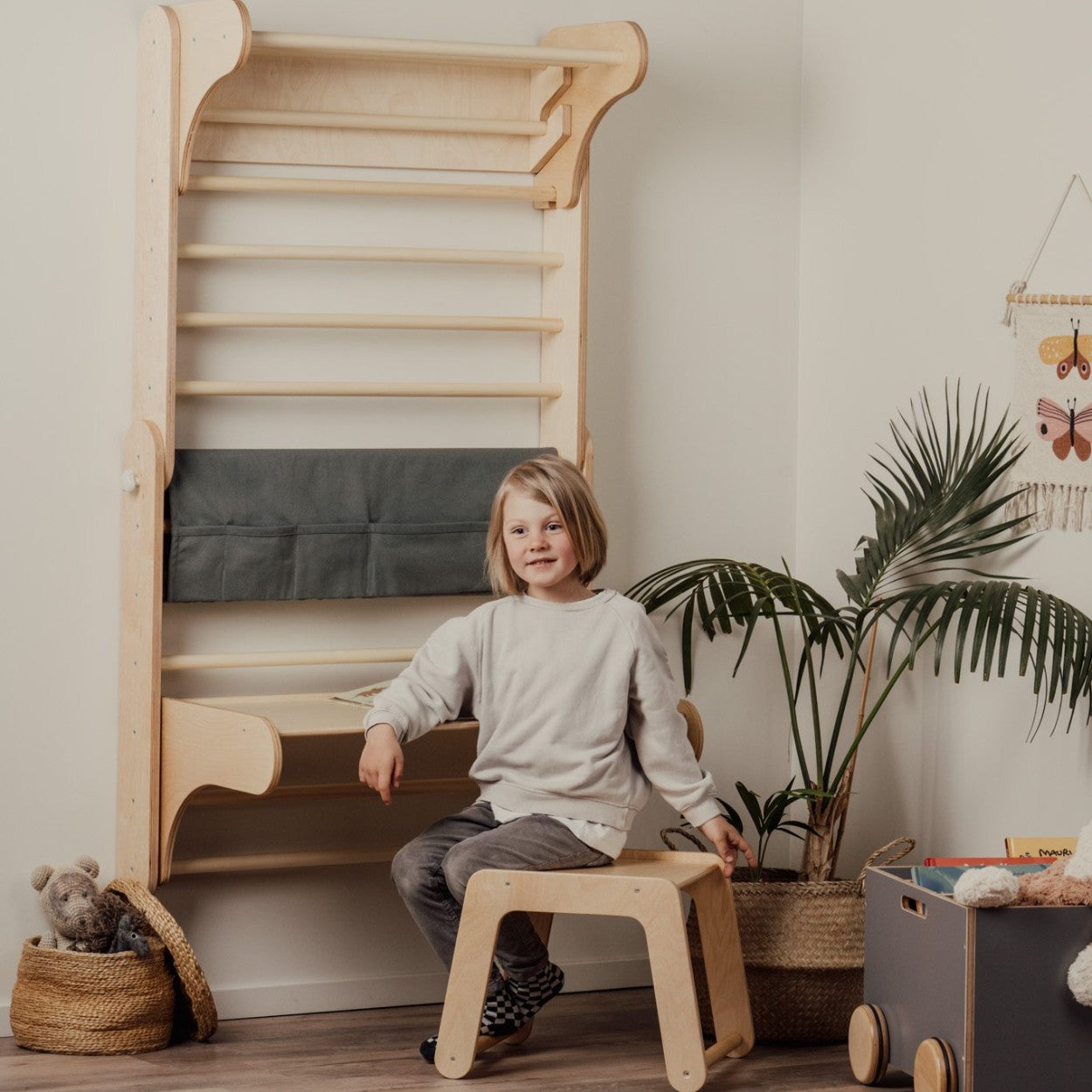 Young child sitting on wooden stool in front of Swedish wall with cushioned mat attachment