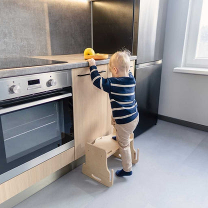 Child reaching up to stovetop while standing on wooden kitchen learning tower