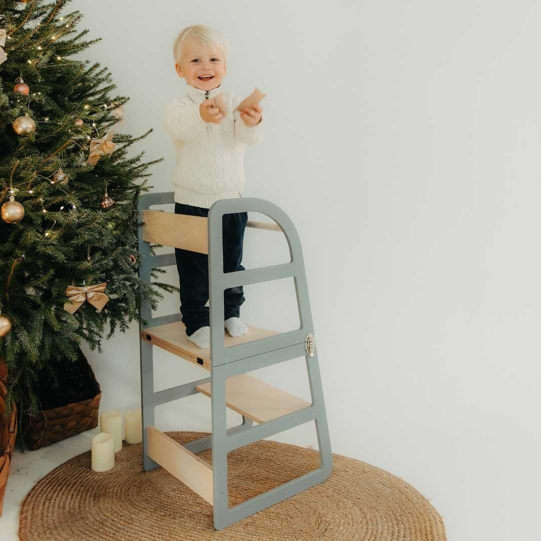 Toddler standing in grey kitchen tower next to decorated Christmas tree
