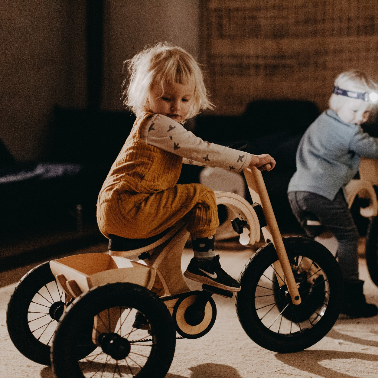 Toddler steering wooden tricycle add-on indoors with another child nearby
