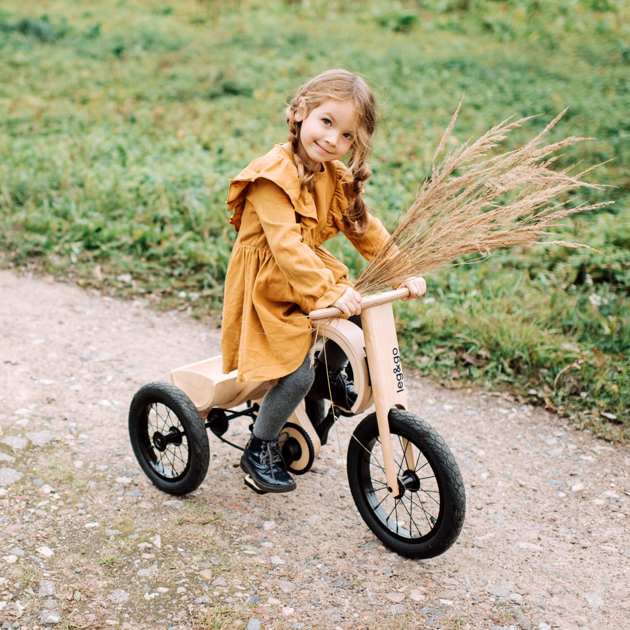 Child in mustard dress riding tricycle add-on on gravel path outdoors