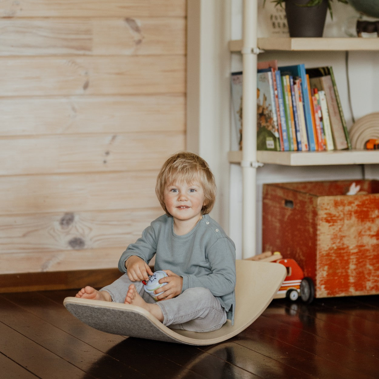 Small child sitting on wooden balance board with grey felt playing with toys