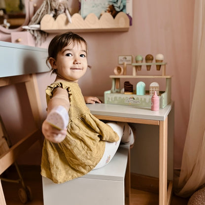 Child in yellow dress playing with wooden ice cream shop set on white desk in bedroom